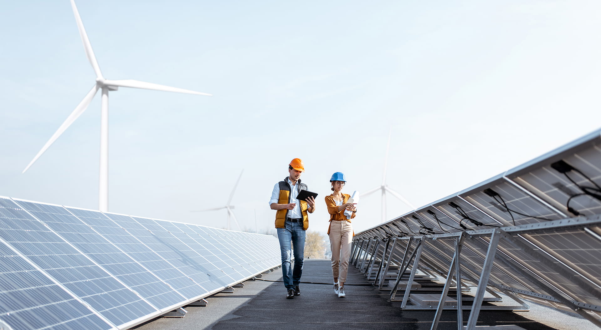 En man och kvinna i skyddsutrustning inspekterar solpaneler. I bakgrunden syns vindkraftverk.